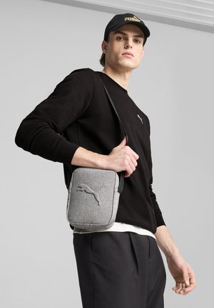 Grå stof crossbody taske med et Puma-logo, rektangulær form, justerbar rem og en struktureret overflade, båret af en model i afslappet tøj.