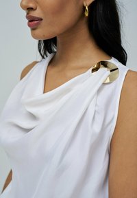 Top de tela blanca con escote drapeado, que presenta un acento circular dorado en el hombro. Textura suave, diseño sin mangas, estilo minimalista.