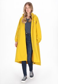 Sárga vízálló esőkabát kapucnival, hosszú ujjakkal és bő szabással. Csíkos tengerészkék felső és kék farmer fölött viselve. Szürke cipő.