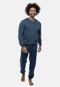 Man met krullend haar die glimlacht, blootsvoets loopt, gekleed in een langärmige donkerblauwe streepjespull en bijpassende broek.