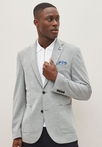 Blazer gris texturé avec deux poches devant, boutons noirs et col cranté. Porté sur un polo blanc zippé, accompagné d'une pochette à motif.