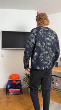 Tamsiai mėlynas megztinis su tekstūruotu raštu, puošiamas šviesesniais gėlių akcentais. Derinamas su juodomis kelnėmis, spalvinga kėde ir šalia esančiu augalu.