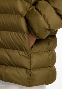Veste en duvet vert olive avec une texture matelassée, des coutures apparentes et un ourlet arrondi. Poche latérale visible, en tissu lisse et brillant.
