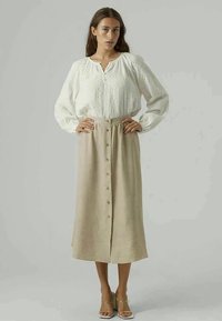 Blouse blanche texturée avec des manches bouffantes, associée à une jupe midi beige clair à boutons sur le devant, créant un look superposé.