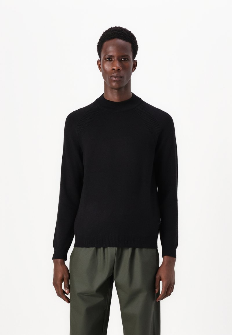 Schwarzer Strickpullover mit hohem Kragen, gerippten Details an den Schultern und langen Ärmeln, kombiniert mit olivgrünen Hosen.
