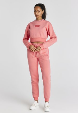 Rosa cropped sweatshirt med logo-lap, parret med matchende joggers med elastisk talje og manchetter. Hvide sneakers fuldender looket.