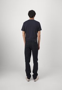 Muž oblečený v čiernom tričku, tmavých džínsoch a bielych teniskách stojí otočený k jednoduchej sivej stene s rukami uvoľnene po bokoch.