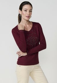 Burgundi hosszú ujjú bordázott pulóver V nyakkal, amely geometrikus mintázatú díszítésekkel rendelkezik az elülső részen.