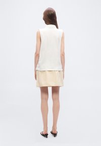 Γυναίκα που στέκεται με την πλάτη, φοράει λευκό αμάνικο τοπ, μπεζ φούστα και μαύρα σανδάλια με τακούνι, μπροστά σε απλό ανοιχτό φόντο.