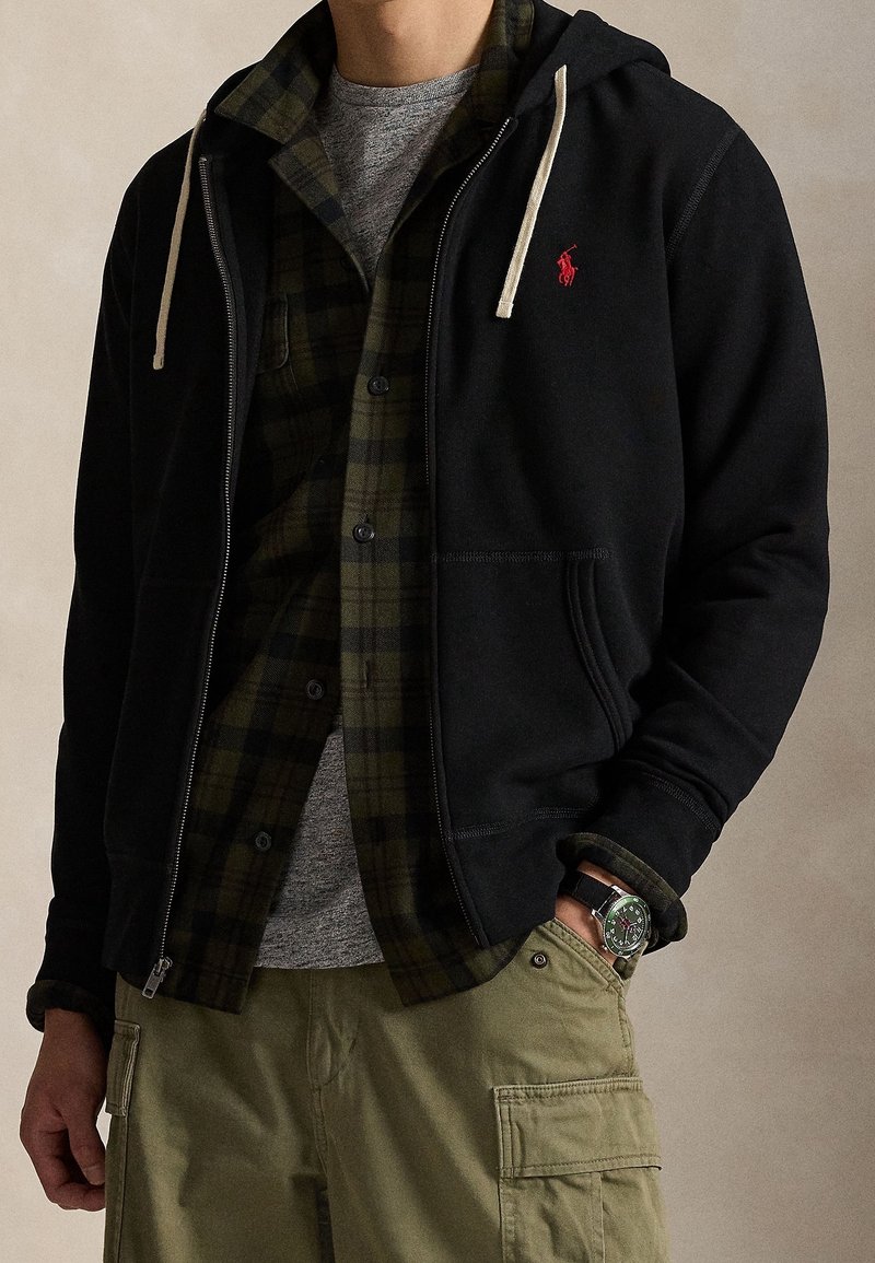 Homme portant des vêtements superposés : sweat à capuche noir zippé avec cordons blancs, chemise à carreaux verts et noirs, t-shirt gris, pantalon cargo vert, montre-bracelet.