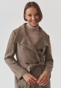 Bruine geruite trenchcoat met een brede kraag, dubbele knopen en een riem. Draag het over een lichte coltrui. Soepele textuur.