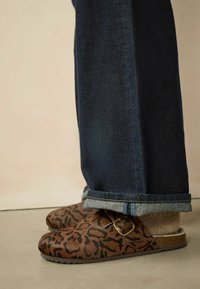 Sabots en cuir marron avec un motif léopard noir, dotés d'une boucle en or, associés à un jean en denim bleu foncé retroussé au niveau de l'ourlet.