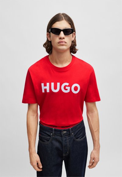 Червена памучна тениска с бял текст "HUGO", съчетана с тъмносини дънкови панталони, носена с черни овнеобразни слънчеви очила. Прост дизайн, удобна кройка.