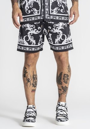 Homme portant un short à motifs noir et blanc, des baskets assorties, avec des tatouages visibles sur les jambes comprenant une araignée, un chien et un aigle.