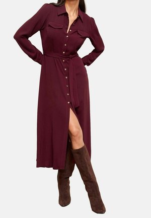 Femme portant une robe bordeaux à manches longues, boutonnée devant, avec des poches poitrine, une ceinture à la taille, une fente frontale et des bottes en daim marron montant jusqu'aux genoux.