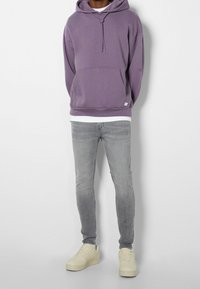 Hombre con sudadera morada, camiseta blanca, jeans skinny desgastados grises y zapatillas beige de pie contra una pared blanca.