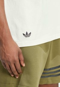 Bílá bavlněná trička s malým černým logem Adidas trefoil poblíž lemu, spárovaná s olivově zelenými šortkami, které mají kontrastní pruhy.
