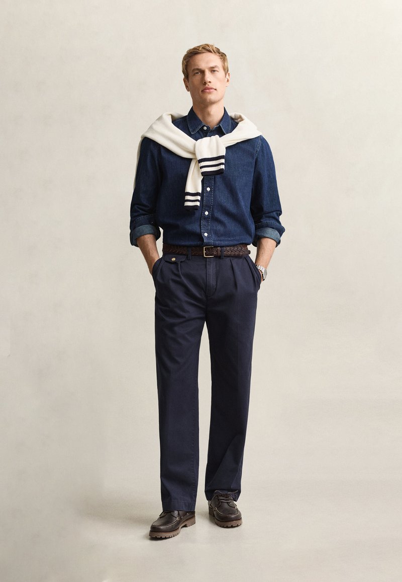 Uomo in piedi con le mani in tasca, indossa una camicia blu scuro, pantaloni blu navy, cintura e scarpe marroni, con un maglione bianco drappeggiato sulle spalle.
