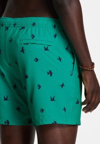 Turquoise swim shorts with a bird pattern in navy blue, featuring an elastic waistband and a rear pocket. Smooth fabric texture.