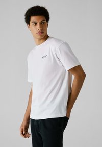 Camiseta de algodón blanca con cuello redondo, mangas cortas y un pequeño logo negro de "Red Bull Racing" en el lado izquierdo del pecho.