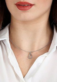 Sølv halskæde med en dråbevedhæng, der har et struktureret design. Båret over en hvid kravebluse, hviler på kravebenet.