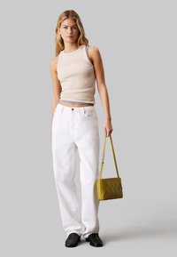Top a maniche lunghe beige a righe con logo Calvin Klein, jeans bianchi larghi, scarpe nere e una borsa a tracolla gialla con dettagli a zigzag.