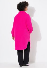 Abrigo largo y esponjoso de color rosa brillante con cuello, aberturas laterales y mangas sueltas. Usado sobre pantalones negros y zapatos de color claro.