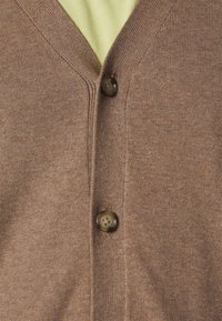 Hnědý kardigan vyrobený z jemně strukturované tkaniny, s véčkovým výstřihem, dvěma hnědými knoflíky a hladkou texturou, vrstvený přes světle zelenou košili.