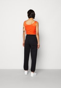 Vrouw met kort donker haar, gekleed in een oranje mouwloos topje, zwarte joggerbroeken, witte sneakers en een ketting, staande tegen een neutrale muur.
