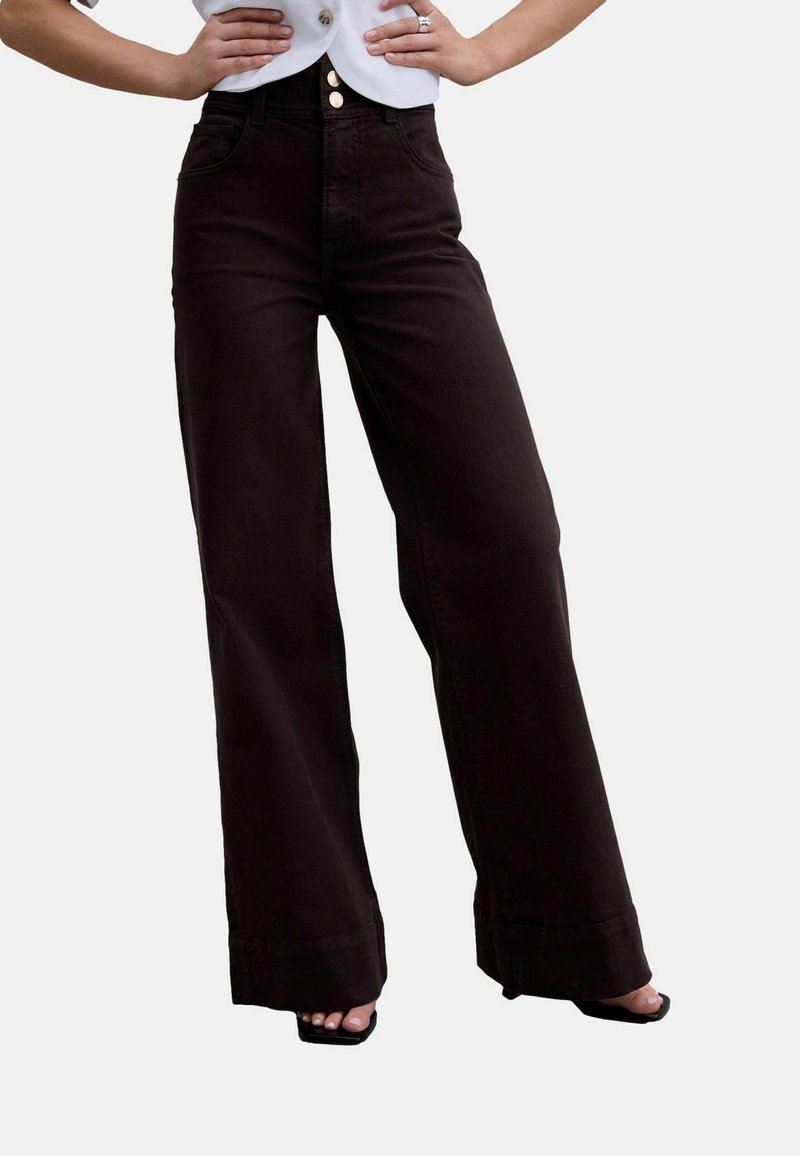 Femme portant un pantalon large taille haute marron foncé avec fermeture à trois boutons dorés, mains sur les hanches, associé à des escarpins ouverts noirs.
