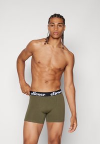 Olivgrüne Boxershorts mit einem schwarzen Bund, der das weiße "ellesse"-Logo trägt. Der Stoff wirkt glatt und enganliegend. Das Model posiert mit einer Hand am Bund.