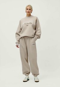Sweatshirt bege com texto de logotipo e calças de treino ajustadas a condizer, ambas em tecido macio. Ténis brancos com pormenores em verde.