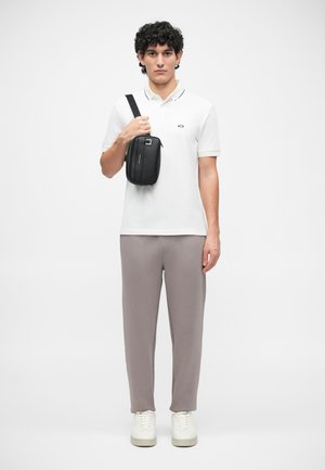 Bílá polo triko s černými akcenty, šedé tepláky a bílé tenisky. Černá crossbody taška s detaily zipu držena vpředu.