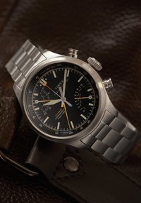 Ασημένιο ρολόι χειρός από ανοξείδωτο ατσάλι με μαύρο καντράν, υποκαντράν χρονογράφου, κλίμακα ταχύμετρου, φωσφορίζοντα δείκτες και πορτοκαλί δευτερεύοντα δείκτη σε σκούρα δερμάτινη επιφάνεια.