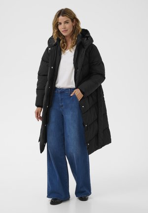 Kvinde iført lang sort pufferjakke med hætte, hvid t-shirt, bredbenede blå jeans og sorte sko, stående mod en hvid baggrund.