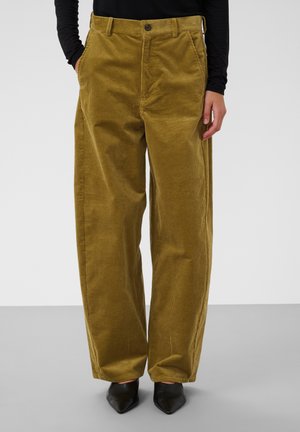 Personne portant un pantalon en velours côtelé vert olive ample avec des poches, associé à des chaussures noires pointues et un haut noir à manches longues.