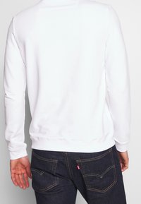 Sudadera blanca de manga larga, con puños acanalados y cuello redondo, combinada con jeans de mezclilla oscura con costuras en contraste.