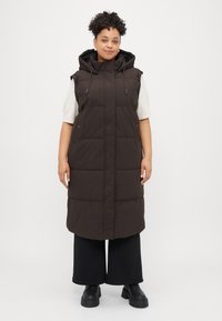 Long, dark brown puffer vest with a hood, featuring a zip closure, side pockets, and a quilted design. Worn over a light top and wide pants.