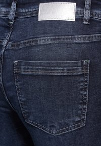 Jean en denim bleu foncé avec une étiquette argentée sur la taille, présentant une poche arrière et des détails de couture contrastants.