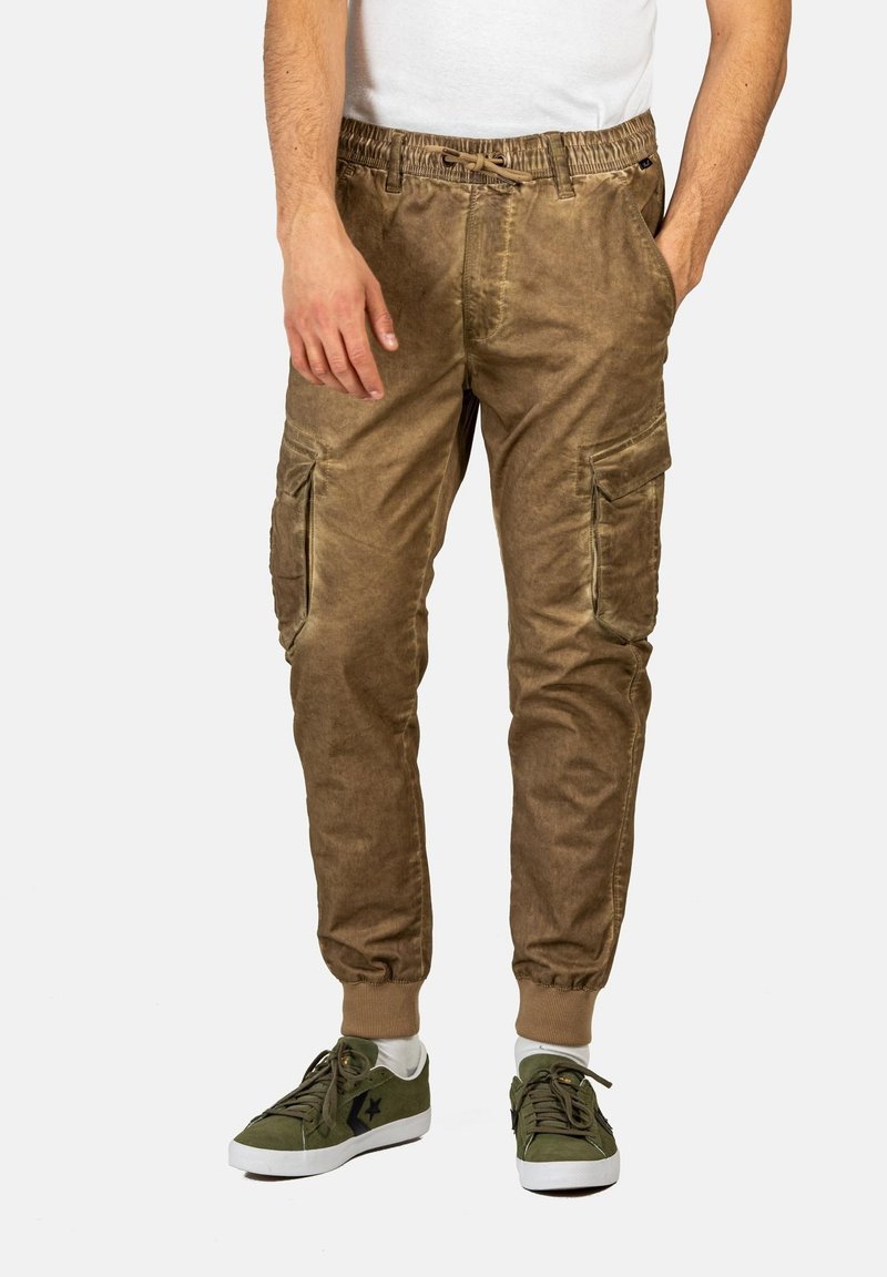 Braune Baumwoll-Cargo-Hosen mit elastischem Bund, seitlichen Taschen und Bündchen, kombiniert mit grünen Turnschuhen. Glatte Textur, lässiges Design.