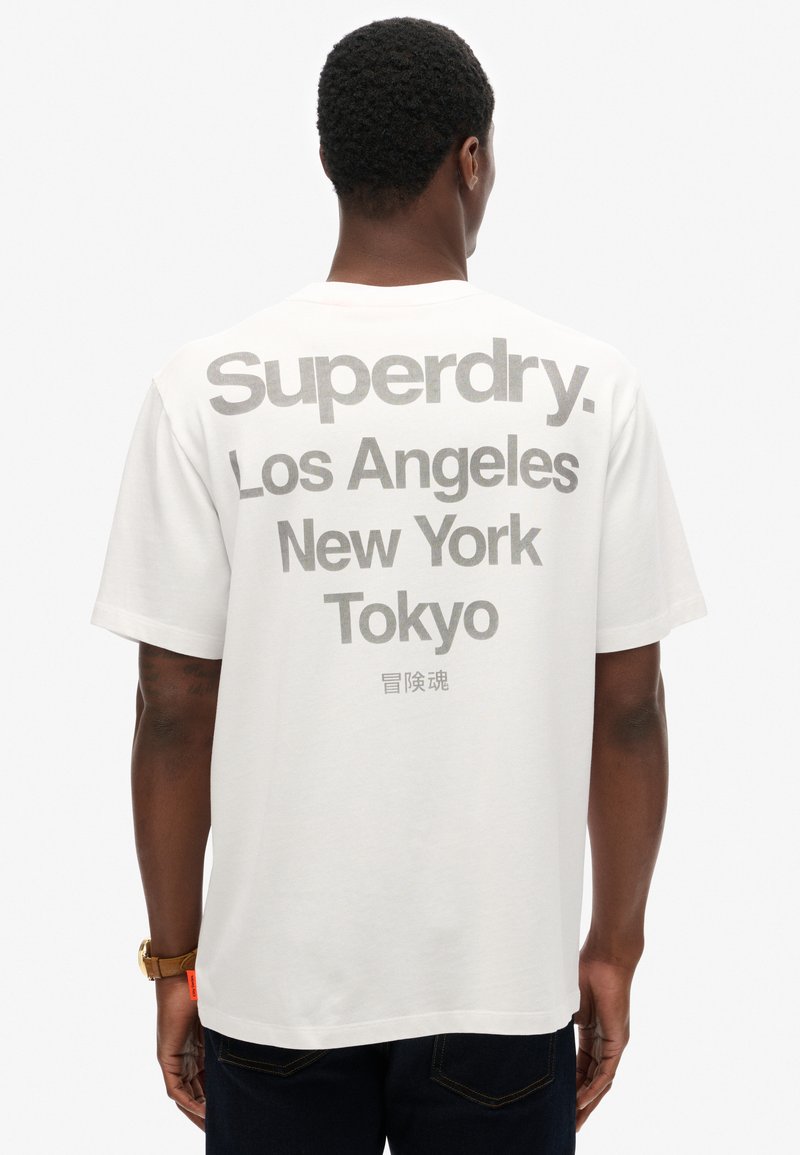 Camiseta blanca de algodón de manga corta, con un gran texto gris en la parte posterior que dice "Superdry," "Los Ángeles," "Nueva York" y "Tokio."