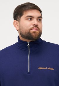 Sudadera azul marino de media cremallera hecha de una tela suave, con un cuello alto y texto bordado en naranja "Originals Studio" en el pecho.
