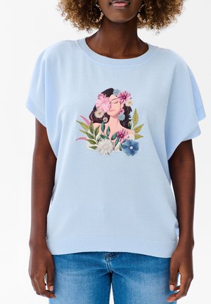 Osoba nosząca jasnoniebieską bluzkę z krótkim rękawem z kwiatowym i kobiecym motywem z przodu, w połączeniu z niebieskimi dżinsami na białym tle.