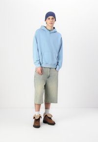Sweatshirt cu glugă de culoare albastru deschis, cu buzunar frontal, asociat cu pantaloni scurți din denim de culoare verde deschis. Botine maro cu accente bej și șosete albe.
