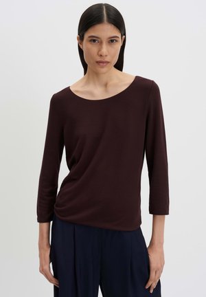 Vrouw met steil donker haar die een donker bordeauxrood shirt met lange mouwen en donkerblauwe broek draagt tegen een effen lichte achtergrond.