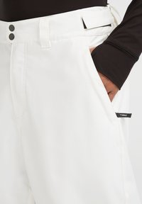 Pantalones blancos con una textura suave, dos cierres de botón negro y un bolsillo lateral con una etiqueta de marca visible.