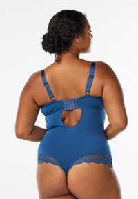 Kvinde med krøllet hår iført en bodysuit med blå blondekanter, der viser justerbare stropper og en lukning bagpå med en nøglehulsknipp.