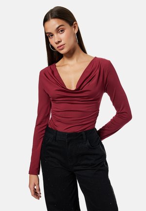 Kobieta o długich ciemnych włosach, nosząca srebrne kolczyki koła, bordową bluzkę z długim rękawem i marszczonym dekoltem oraz czarne spodnie z wysokim stanem.