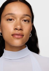 Zbliżenie osoby o brązowych oczach i gładkiej skórze, ubranej w jasnoniebieski top z wysokim kołnierzem i srebrne kolczyki w kształcie koła. Widoczne czarne, falowane włosy.