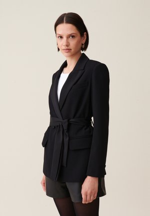 Blazer noir avec une taille ajustée et une ceinture, comportant deux poches avant, un col en V et une texture lisse. Porté par-dessus une chemise blanche.